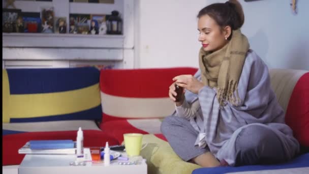 Vista lateral una mujer joven se siente mal. Una joven sentada en un sofá con una bufanda bebe medicina. — Vídeo de stock
