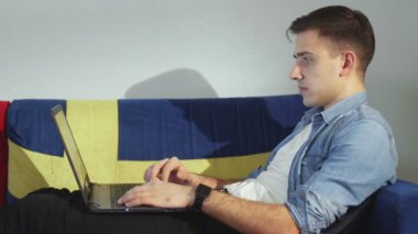 Yan bakışta genç bir adam kanepede yatar ve dizüstü bilgisayarla çalışır, klavyeye yazar, evden uzaktan kumandayla çalışır..