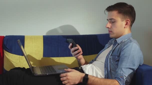Vista lateral un hombre joven está acostado en el sofá y trabajando en un ordenador portátil, escribiendo en el teclado, trabajo remoto desde casa. — Vídeos de Stock