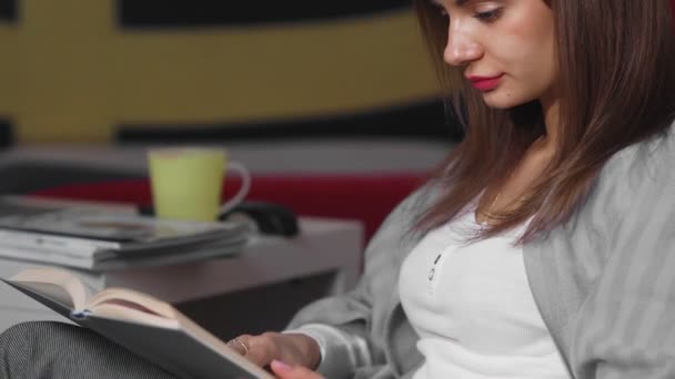 Eine junge Frau sitzt zu Hause auf einem Hocker und liest ein Buch. Eine Frau ruht sich in ihrer Freizeit auf einem bequemen Hocker aus. Schöne freie Zeit am Wochenende — Stockvideo