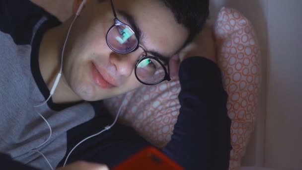 한 젊은 남자가 헤드폰을 끼고 침대에 누워 밤에 인터넷을 보고 있는 모습. — 비디오