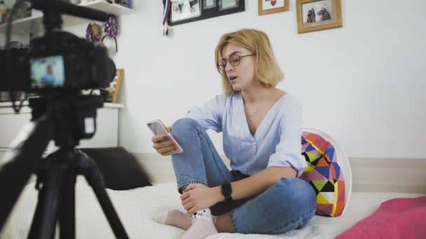 Een jonge vrouw met een bril zit op de bank te glimlachen, pratend met de camera — Stockvideo