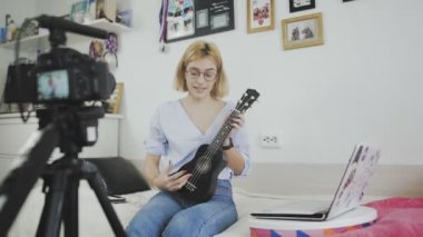 Gözlüklü genç bir kadın kanepede oturuyor, gülümsüyor ve kameraya ukulele çalıyor. Ukulele çalıyorum. Video Blogçusu