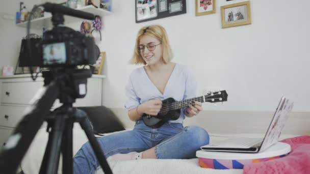 Молодая женщина в очках сидит на диване, улыбается и играет на укулеле перед камерой. Играю на укулеле. Видеоблогер — стоковое видео