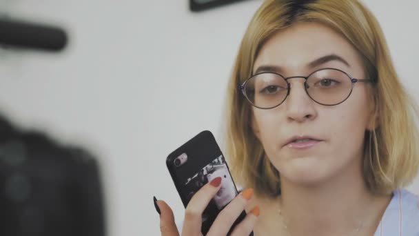 Primer plano de una joven con gafas sentada con un teléfono en las manos, sonriendo y hablando con la cámara — Vídeos de Stock