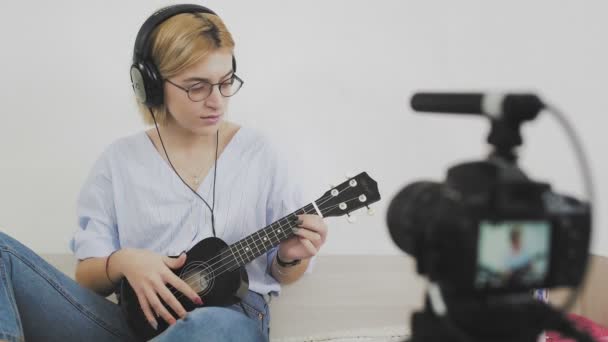 Młoda kobieta w okularach siedzi na kanapie, uśmiecha się i gra na ukulele do kamery. Gram na ukulele. Blogger wideo — Wideo stockowe