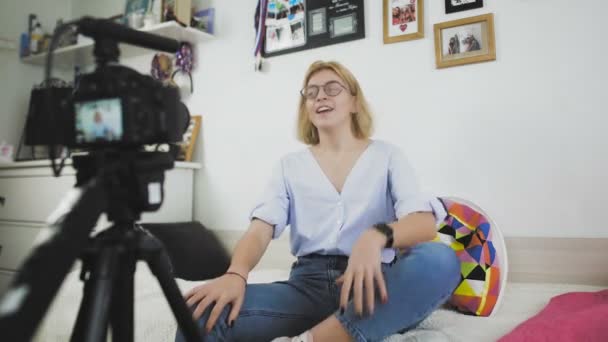 Uma jovem com óculos está sentada no sofá sorrindo, conversando com a câmera — Vídeo de Stock