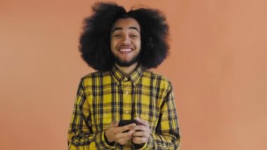 Turuncu arka planda Afrikalı saç stili olan genç bir adam telefona bakar ve mutlu olur. Renkli bir arkaplanda duygular