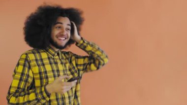 Turuncu arka planda Afrikalı saç stili olan genç bir adam telefona bakar ve mutlu bir şekilde şaşırır. Renkli bir arkaplanda duygular