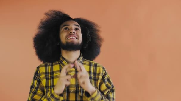 Egy afrikai frizurájú, narancssárga hátterű fiatalember keresztbe teszi az ujjait és imádkozik. Érzelmek egy színes háttér. — Stock videók