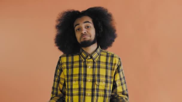 Een jonge man met een Afrikaans kapsel op een oranje achtergrond haalt zijn schouders op. Emoties op een gekleurde achtergrond. — Stockvideo