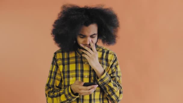 Młody mężczyzna z afrykańską fryzurą na pomarańczowym tle patrzy na telefon i jest szczęśliwie zaskoczony. Emocje na kolorowym tle — Wideo stockowe