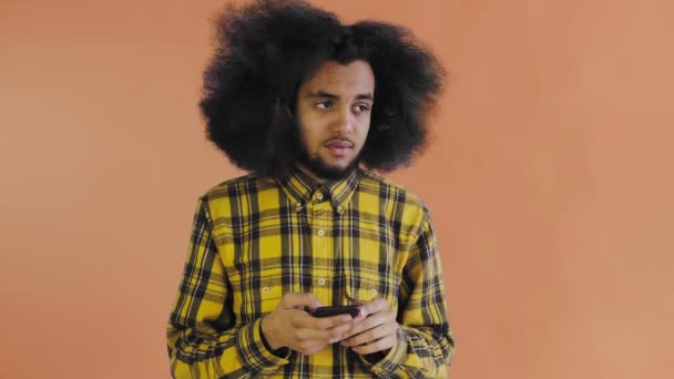 Ένας νεαρός με αφρικάνικο χτένισμα σε πορτοκαλί φόντο κοιτάζει το τηλέφωνο και αμφιβάλλει. Συναισθήματα σε έγχρωμο φόντο — Αρχείο Βίντεο