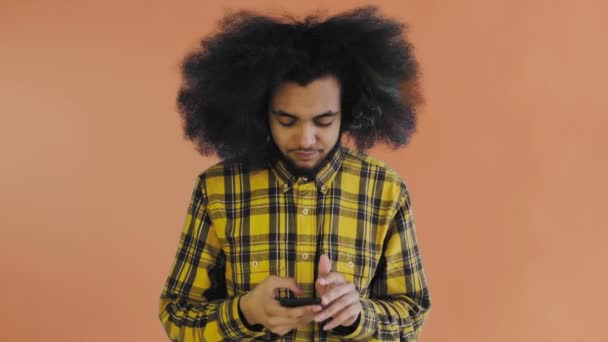 Молодой человек с африканской прической на оранжевом фоне вытаскивает свой телефон, но он не работает. Эмоции на цветном фоне — стоковое видео
