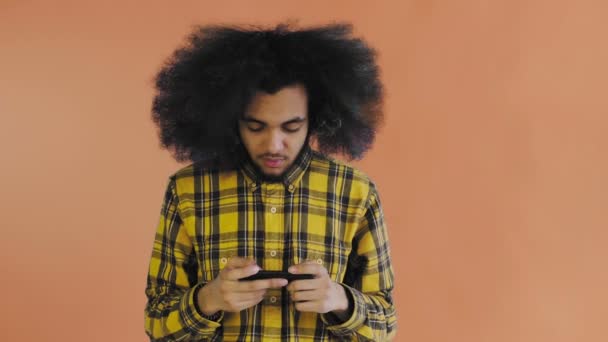 Un joven con un peinado africano sobre un fondo naranja está jugando por teléfono. Sobre un fondo de color — Vídeos de Stock