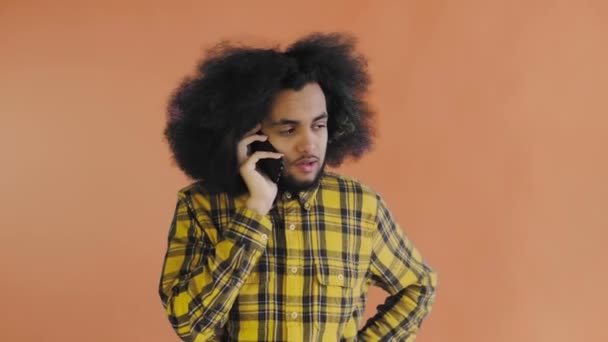 En ung man med en afrikansk frisyr på orange bakgrund pratar i telefon. På en färgad bakgrund — Stockvideo