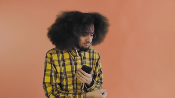 Seorang pemuda dengan gaya rambut Afrika pada latar belakang oranye mendengarkan musik dan headphone. Pada latar belakang berwarna — Stok Video