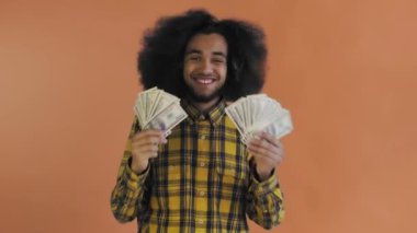 Elinde banknot yelpazesi olan mutlu bir Afro-Amerikalı adam, turuncu bir arka planda izole bir şekilde kameraya bakıyor.