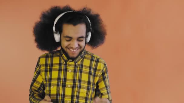 En ung man med afrikansk frisyr på orange bakgrund lyssnar på musik och hörlurar. På en färgad bakgrund — Stockvideo