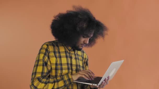 Un joven con un peinado africano sobre un fondo naranja está escribiendo en un portátil. Sobre un fondo de color — Vídeos de Stock