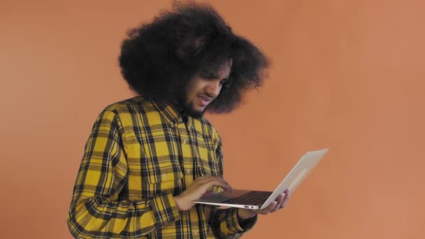 Un joven con un peinado africano sobre un fondo naranja está escribiendo en un portátil. Sobre un fondo de color — Vídeos de Stock