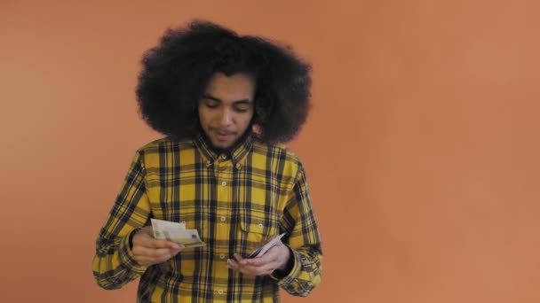 Ένας ευτυχισμένος Αφροαμερικανός που κρατάει χαρτονομίσματα στα χέρια του, τα μετράει και κοιτάζει την κάμερα, στέκεται απομονωμένος σε πορτοκαλί φόντο. — Αρχείο Βίντεο