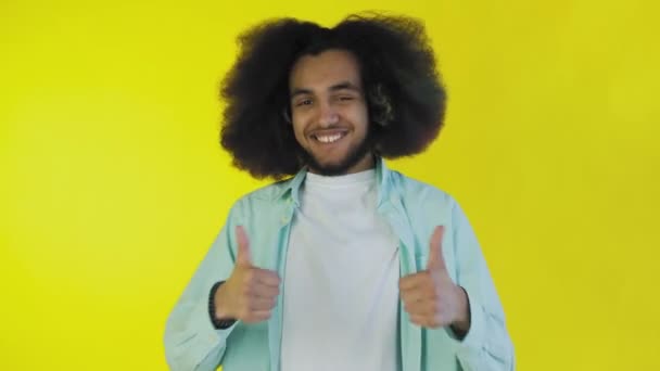 En glad afroamerikansk man tittar in i kameran, visar en liknande med handen, står isolerad på en gul bakgrund — Stockvideo