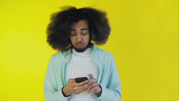 En ung man med en afrikansk frisyr på orange bakgrund tittar på telefonen och är glad. Känslor på en färgad bakgrund — Stockvideo