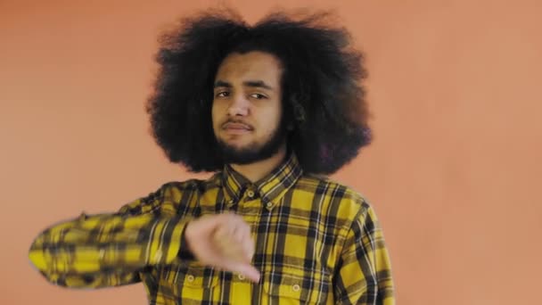 한 흑인 남자가 카메라를 쳐다보고, 엄지손가락을 내리고, 오렌지 색 배경 위에 고립된 채서 있습니다. — 비디오