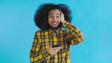 Mavi arka planda Afrikalı saç stili olan genç bir adam telefona bakar ve mutlu bir şekilde şaşırır. Renkli bir arkaplanda duygular