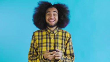 Mavi arka planda Afrikalı saç stili olan genç bir adam telefona bakar ve mutludur. Renkli bir arkaplanda duygular