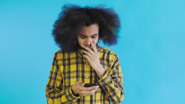 Un joven con un peinado africano sobre un fondo azul mira el teléfono y está felizmente sorprendido. Emociones sobre un fondo coloreado — Vídeos de Stock