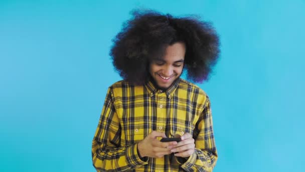 A young man with an African hairstyle on a blue background looks at the phone and happily texts with someone. Emotions on a colored background — Stock Video
