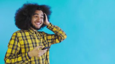 Mavi arka planda Afrikalı saç stili olan genç bir adam telefona bakar ve mutlu bir şekilde şaşırır. Renkli bir arkaplanda duygular