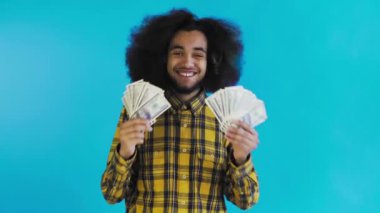 Elinde banknot yelpazesi olan mutlu bir Afro-Amerikalı adam mavi bir arka planda tek başına kameraya bakıyor.