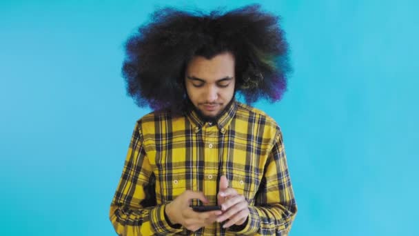 Молодой человек с африканской прической на синем фоне вытаскивает свой телефон, но он не работает. Эмоции на цветном фоне — стоковое видео