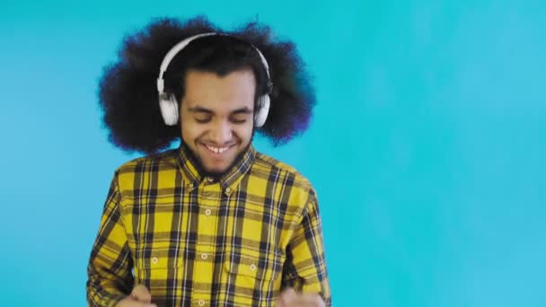 En ung man med en afrikansk frisyr på blå bakgrund lyssnar på musik och hörlurar. På en färgad bakgrund — Stockvideo