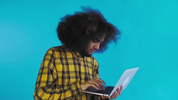 Un joven con un peinado africano sobre un fondo azul está escribiendo en un portátil. Sobre un fondo de color — Vídeos de Stock