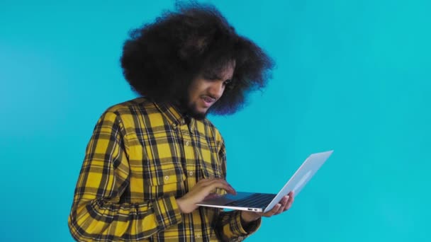 Un joven con un peinado africano sobre un fondo azul está escribiendo en un portátil. Sobre un fondo de color — Vídeos de Stock