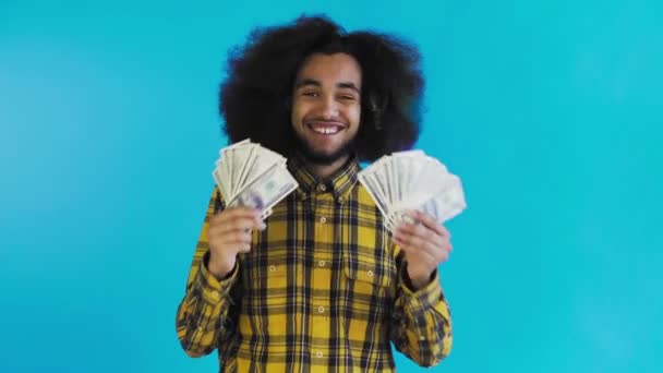 青い背景に孤立して立っているカメラを見て、彼の手に紙幣のファンを保持し、幸せなアフリカ系アメリカ人の男 — ストック動画