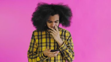 Pembe arka planda Afrikalı saç stili olan genç bir adam telefona bakar ve mutlu bir şekilde şaşırır. Renkli bir arkaplanda duygular