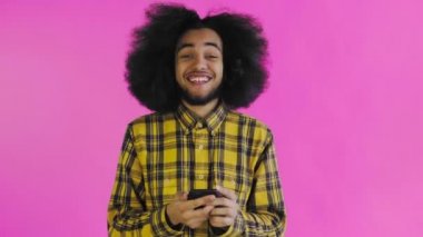 Pembe arka planda Afrikalı saç stili olan genç bir adam telefona bakar ve mutludur. Renkli bir arkaplanda duygular