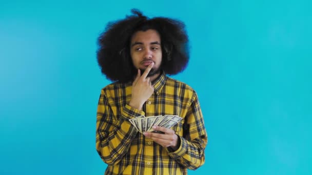 Um afro-americano feliz segurando dinheiro em suas mãos e pensando sobre o que gastá-lo, fica isolado em um fundo azul — Vídeo de Stock