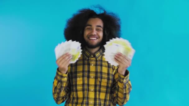 Un hombre afroamericano feliz sosteniendo un abanico de billetes en sus manos y mirando a la cámara, parado aislado sobre un fondo azul — Vídeo de stock