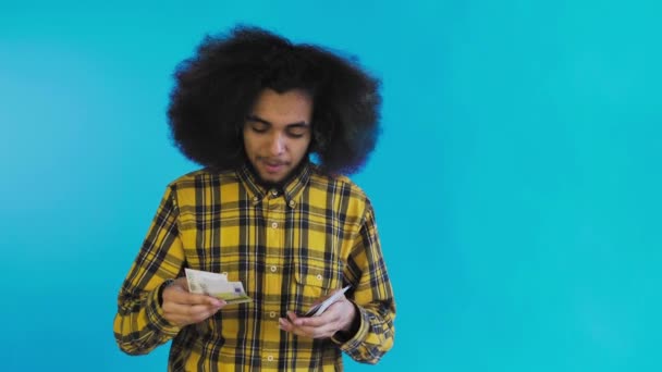 Seorang pria Afrika-Amerika yang bahagia memegang uang kertas di tangannya, menghitung mereka dan melihat ke kamera, berdiri terisolasi di latar belakang biru — Stok Video