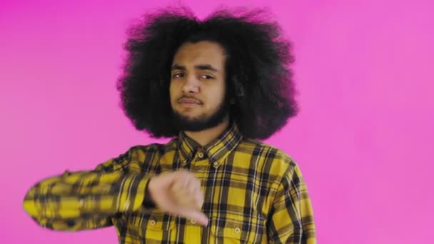 Афроамериканец смотрит в камеру, опускает пальцы, стоит изолированный на розовом фоне — стоковое видео