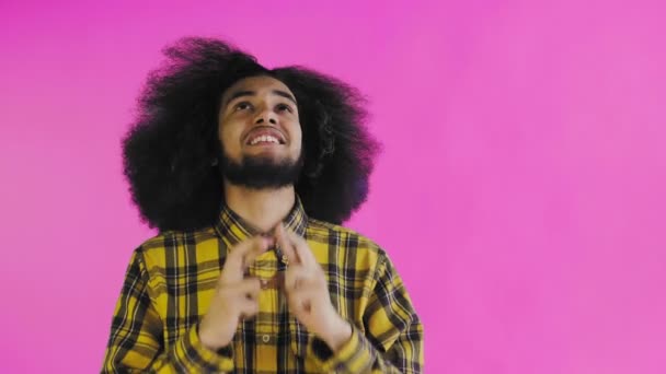 Egy afrikai frizurájú, rózsaszín hátterű fiatalember keresztbe teszi az ujjait és imádkozik. Érzelmek egy színes háttér. — Stock videók