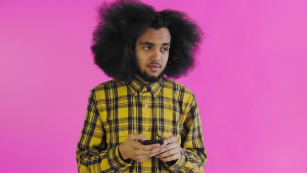 Młody mężczyzna z afrykańską fryzurą na różowym tle patrzy na telefon i ma wątpliwości. Emocje na kolorowym tle — Wideo stockowe