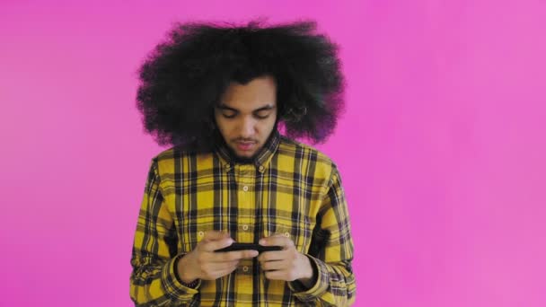 Un giovane con un'acconciatura africana su sfondo rosa sta giocando al telefono. Su uno sfondo colorato — Video Stock