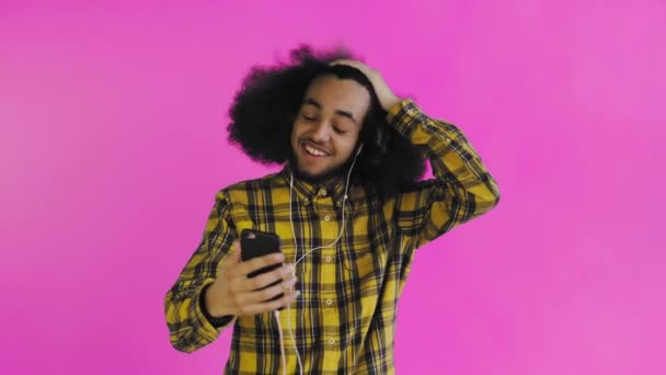 Un joven con un peinado africano sobre un fondo rosa en auriculares se comunica a través de un enlace de vídeo. Sobre un fondo de color — Vídeos de Stock